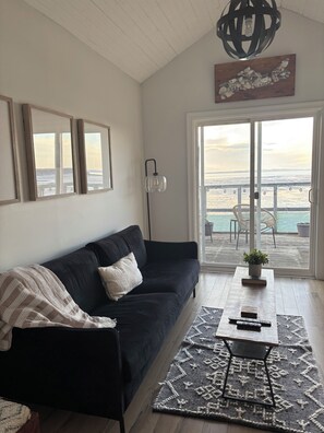 A living room with a black sofa, a coffee table with a plant, and a view of the ocean through a sliding glass door. - Beautiful Oceanfront Home on the Minas Basin (Newport)