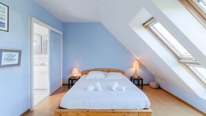 Une chambre avec un plafond en pente, un lit recouvert de draps blancs, deux lampes de chevet et un puits de lumière.
