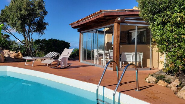 A swimming pool with lounge chairs, a glass-enclosed patio, and a view of the surrounding landscape. - DomusParadiso (Costa Paradiso)