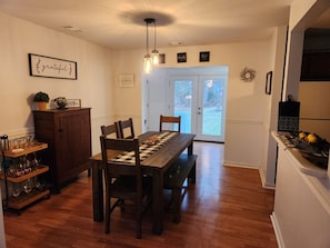 A dining area with a wooden table, chairs, and a chandelier. There is a cabinet with glassware and a potted plant on the left. The room has a white door leading outside.