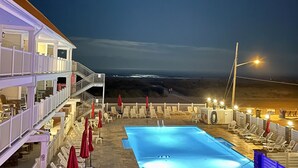 Un área de piscina en la azotea con vista a la playa y al océano, rodeada de sillas de descanso y sombrillas.