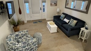 Una sala de estar con un sofá azul oscuro, un sillón estampado, una mesa de centro blanca y una mesita auxiliar. Hay una planta en maceta, un televisor montado en la pared y una puerta con una paleta de madera al fondo.