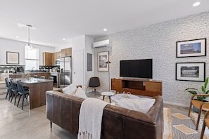 A modern living room with a brown leather sofa, a flat-screen TV, and a kitchen area with wooden cabinets and a dining set. - South Side Modern | DT Chattanooga| A+ Walkable (Chattanooga)