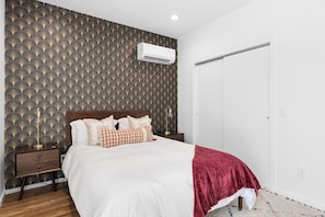 Un dormitorio con papel tapiz estampado, una cama con ropa de cama blanca, un plaid rojo, una mesita de noche y un aire acondicionado instalado en la pared.