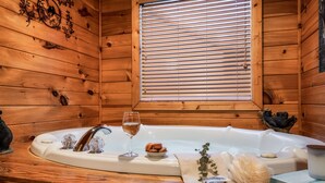 A wooden cabin interior with a large bathtub, a window with blinds, and various toiletries on the side.