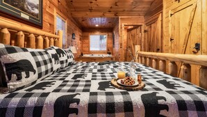 A wooden cabin room with a checkered bedspread, a framed picture on the wall, and a window with blinds.