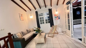 Un salon avec un canapé, une table basse et un ventilateur. Il y a une fenêtre avec des rideaux et une porte qui donne sur l’extérieur.