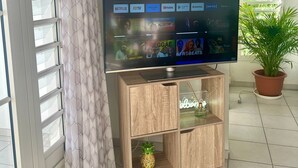 Un téléviseur posé sur un meuble en bois, avec une étagère en verre en dessous, une plante en pot à droite et une fenêtre avec des stores à gauche.