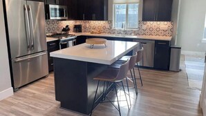 A modern kitchen with dark cabinetry, stainless steel appliances, a central island with bar stools, and a window providing natural light. - Riverstone Village Pet Friendly Beach Cruisers Sleeps 6 (Coeur d'Alene)