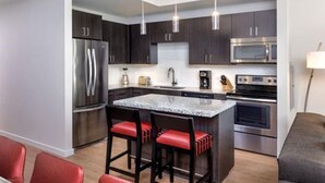 A modern kitchen with dark cabinets, stainless steel appliances, a granite countertop island with red bar stools, and pendant lighting. - 2BD/Austin - Full Kitchen, W/D, Balcony, Fire Pit! (Austin)