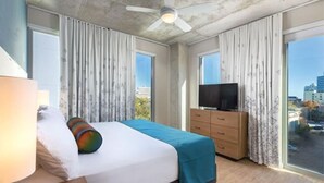 A hotel room with a bed, a television, a dresser, and large windows offering a city view. - 2BD/Austin - Full Kitchen, W/D, Balcony, Fire Pit! (Austin)