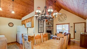 Une cuisine chaleureuse et rustique avec un plafond en bois, un coin repas avec une table et des chaises en bois, une cheminée en pierre et une horloge au mur.