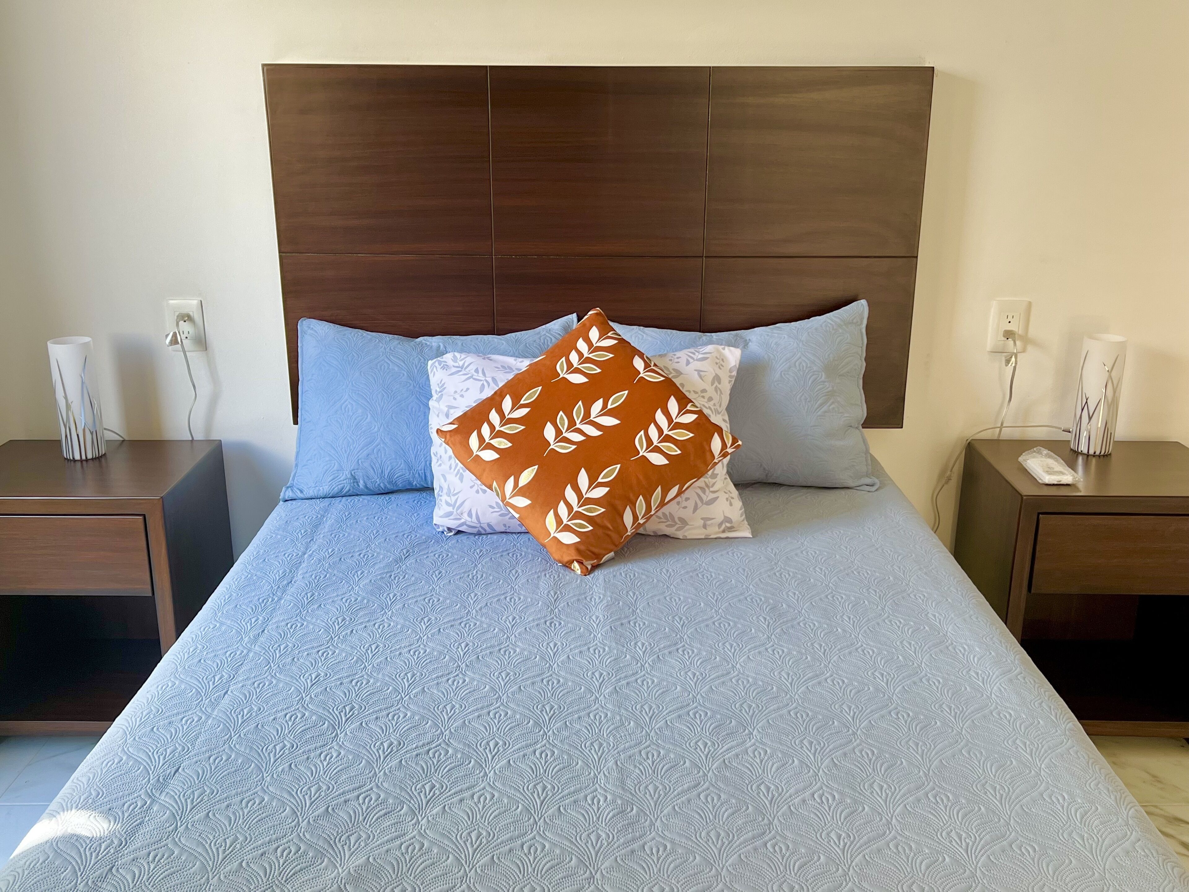 A neatly made bed with a patterned bedspread, a brown leaf-patterned pillow, and two matching nightstands with lamps and phone chargers.