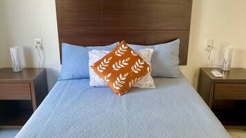 A neatly made bed with a patterned bedspread, a brown leaf-patterned pillow, and two matching nightstands with lamps and phone chargers.