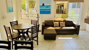 A living room with a dining area, a sofa, and a TV. There are two windows with blinds, a ceiling fan, and a painting on the wall. - Gorgeous cozy apartment with pool, balcony and roof top in Punta Sur! Casa Ama 4 (Isla Mujeres)