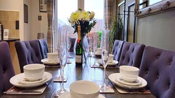Mesa de comedor dispuesta para seis personas, con platos, cuencos y copas de vino blancos, flanqueada por sillas moradas tapizadas. Un jarrón con flores blancas está centrado en la mesa.