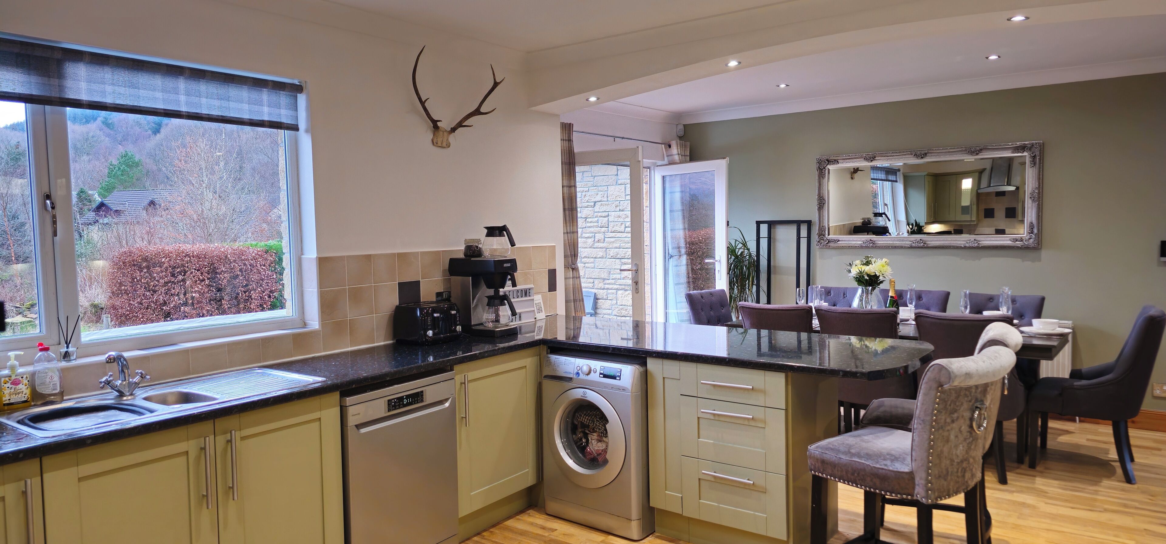 A modern kitchen with a built-in dishwasher, a washing machine, and a coffee maker. There is a dining area with a table and chairs, and a large mirror on the wall.