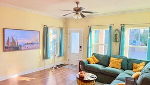 A living room with a sofa, a painting, a ceiling fan, and a window with curtains. - Casa del Sole • Downtown • 3 Ensuites • Pets OK (Pensacola)