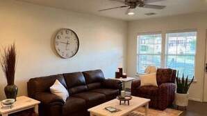 A living room with a brown leather sofa, a white coffee table, a patterned rug, a clock on the wall, and a ceiling fan.