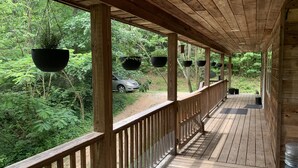 Una terraza de madera con macetas colgantes, un espacio cubierto y vista a un área boscosa con un coche al fondo.