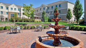 A courtyard with a central fountain, outdoor seating, and a backdrop of multi-story buildings. - 1 Bedroom | Pool & Gym. Free Breakfast (O'Fallon)