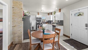 Une cuisine de taille réduite avec des meubles blancs, un mur décoratif en briques et un coin repas comprenant une table et des chaises en bois.