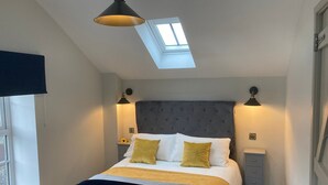 Une chambre à coucher avec un puits de lumière, une tête de lit capitonnée, deux lampes de chevet et un lit recouvert d’une couverture à rayures et de deux oreillers.