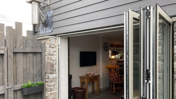 Une entrée de maison agrémentée d’un mur en pierre, d’une clôture en bois et d’une porte-fenêtre coulissante donnant sur un espace de vie comprenant un téléviseur, une table en bois et un fauteuil à bascule.