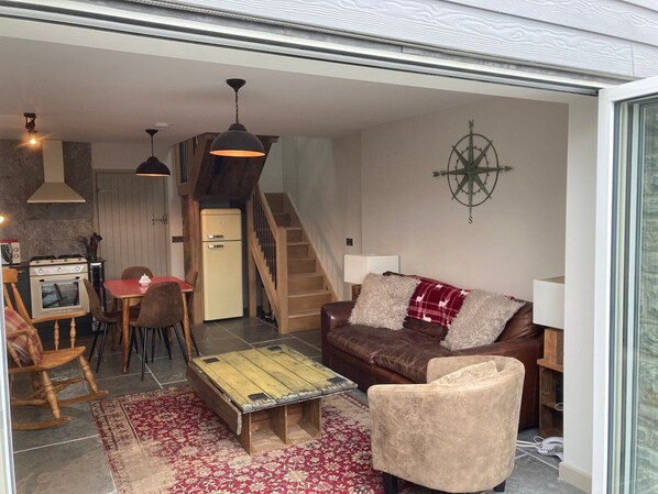 A cozy living room with a brown leather sofa, a wooden coffee table, and a red patterned rug. A wooden rocking chair and a dining table with chairs are also visible.