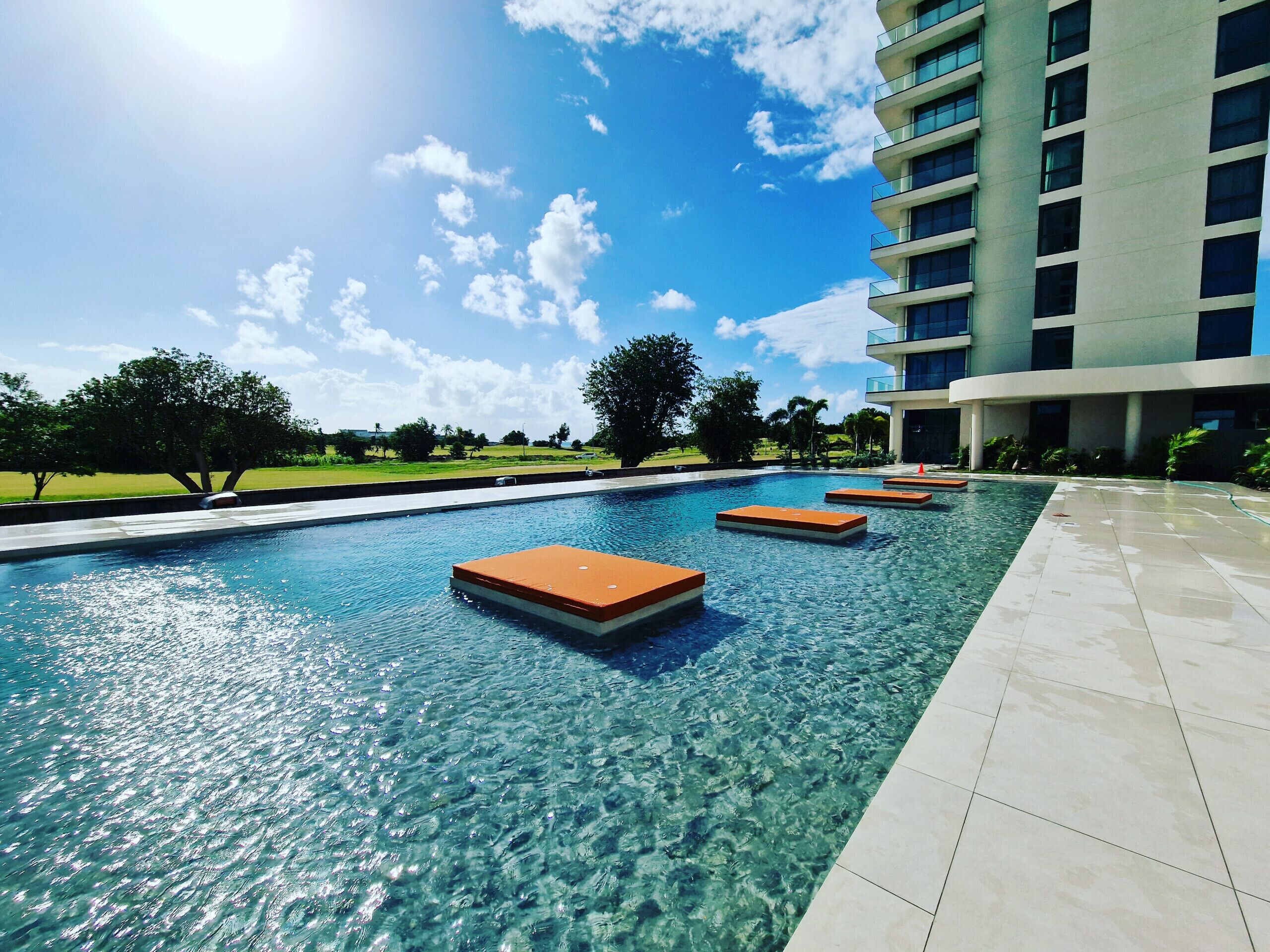 A clear swimming pool with floating platforms, surrounded by a modern building and a grassy area with trees.