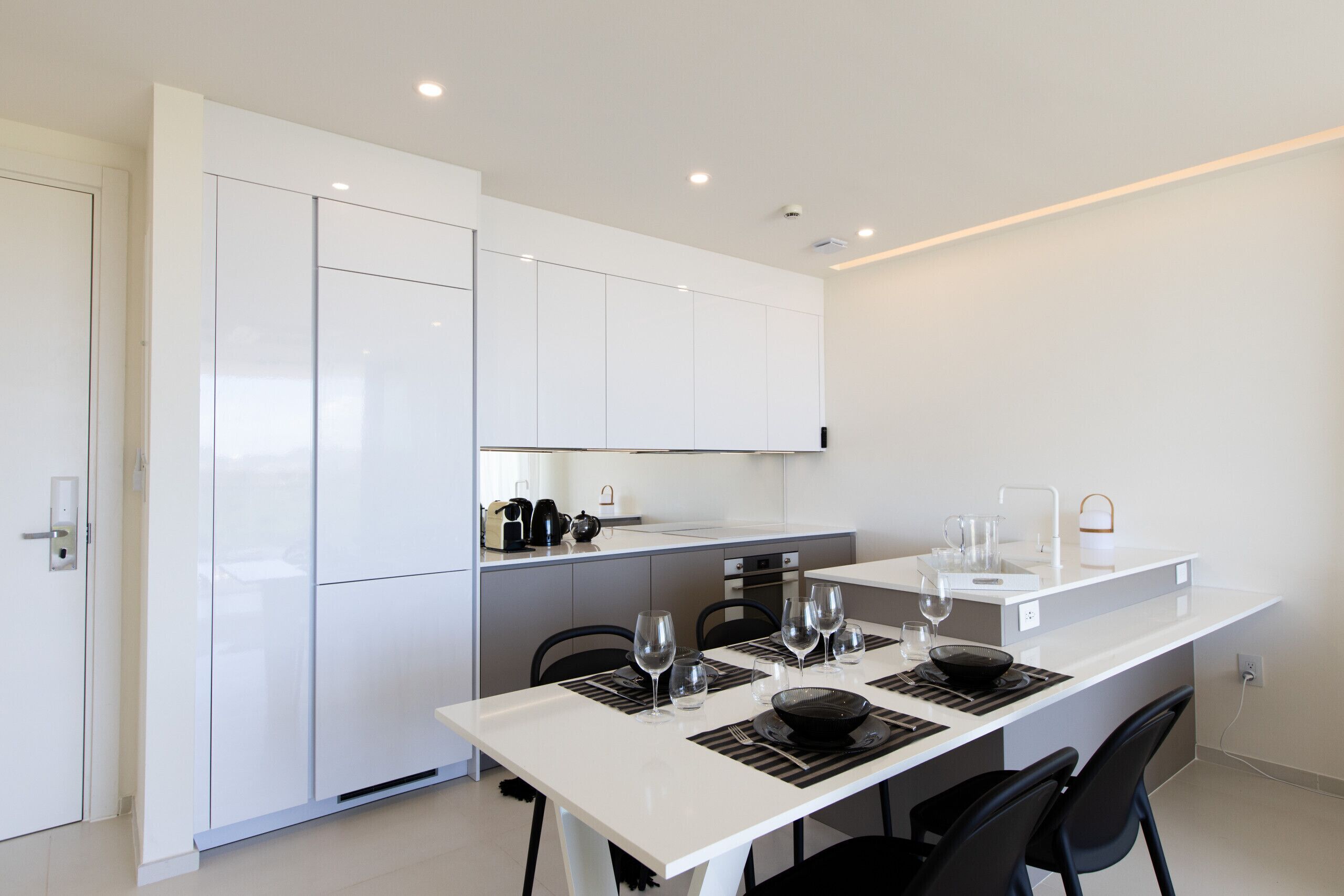 Een moderne keuken met witte kasten, een eettafel gedekt met wijnglazen en een klein keukeneiland met een waterkoker.