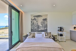 Une chambre dotée d’un grand lit, offrant une vue sur la plage, avec un coin café et un tableau représentant des palmiers.