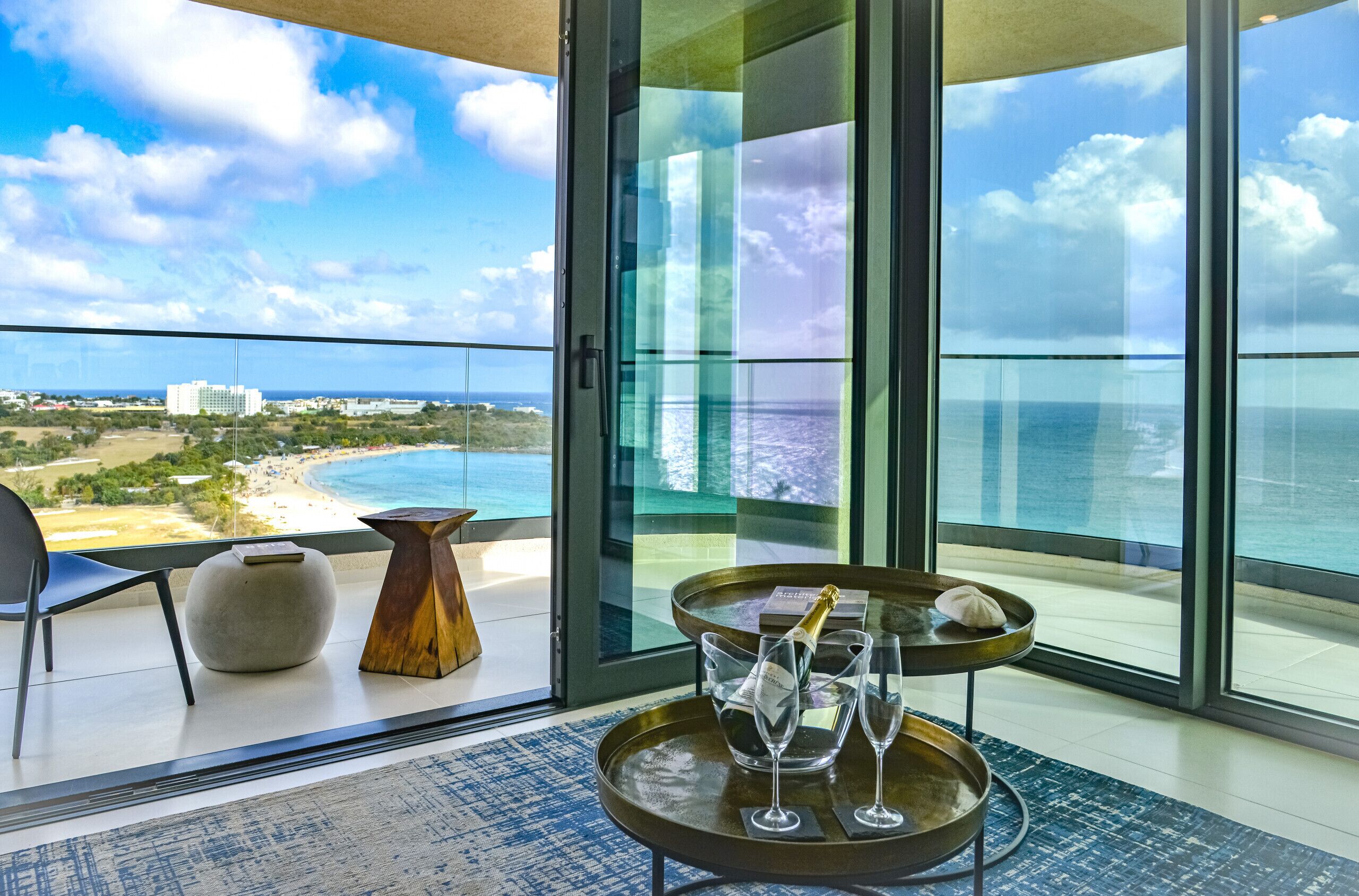 A balcony with a view of the ocean and beach, a table with a bottle of champagne, and a chair.