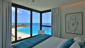 A bedroom with a large window offering a view of a beach and ocean. - A-1701 Panoramic ocean view 2 bedroom apartment (Cupecoy)