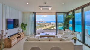 A modern living room with a large flat-screen TV, a wooden entertainment unit, a beige sofa, and a view of the ocean through large glass doors. - A-1701 Panoramic ocean view 2 bedroom apartment (Cupecoy)