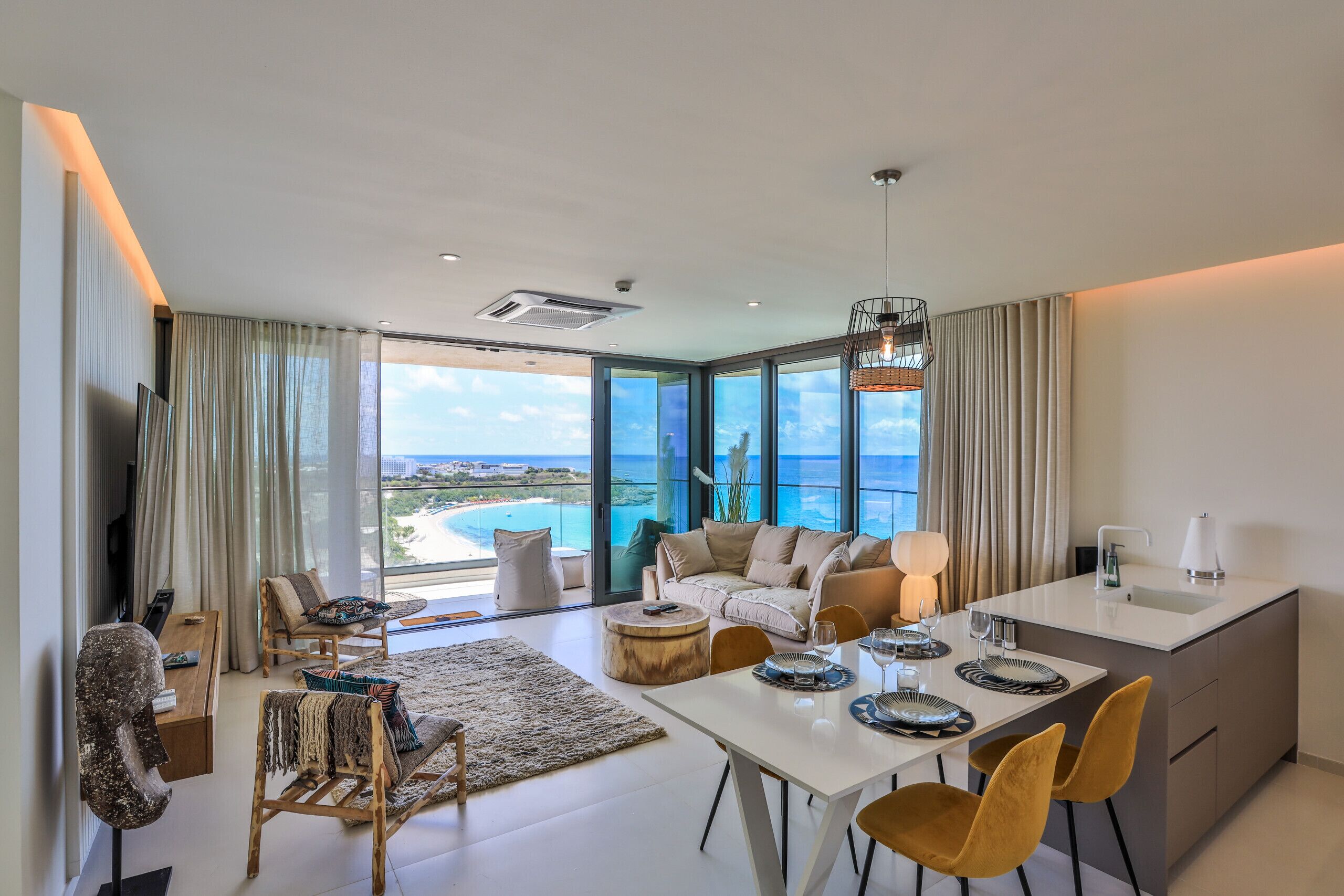 A modern living room with a large window overlooking a beach, a dining area with a white table, and a kitchen with a sink and countertop.