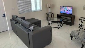 Ein modernes Wohnzimmer mit einer Eckcouch, einem Glastisch, einem Fernseher und einer gemusterten Tischdecke.