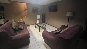 A living room with two purple sofas, a small table, a TV mounted on the wall, and a staircase in the background. - Cozy 2 bedroom suite with private entrance and continental breakfast. (Port Matilda)