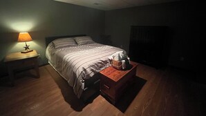 A dimly lit bedroom with a bed, a nightstand with a lamp, and a wooden chest of drawers. - Cozy 2 bedroom suite with private entrance and continental breakfast. (Port Matilda)