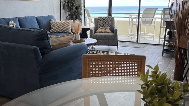 Una sala de estar moderna con un sofá azul, una mesa redonda de vidrio y vistas a la playa a través de amplios ventanales correderas.