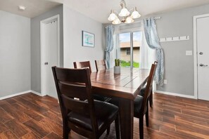 Een eetkamer met een houten tafel en stoelen, een kroonluchter en uitzicht op het groen door grote ramen.