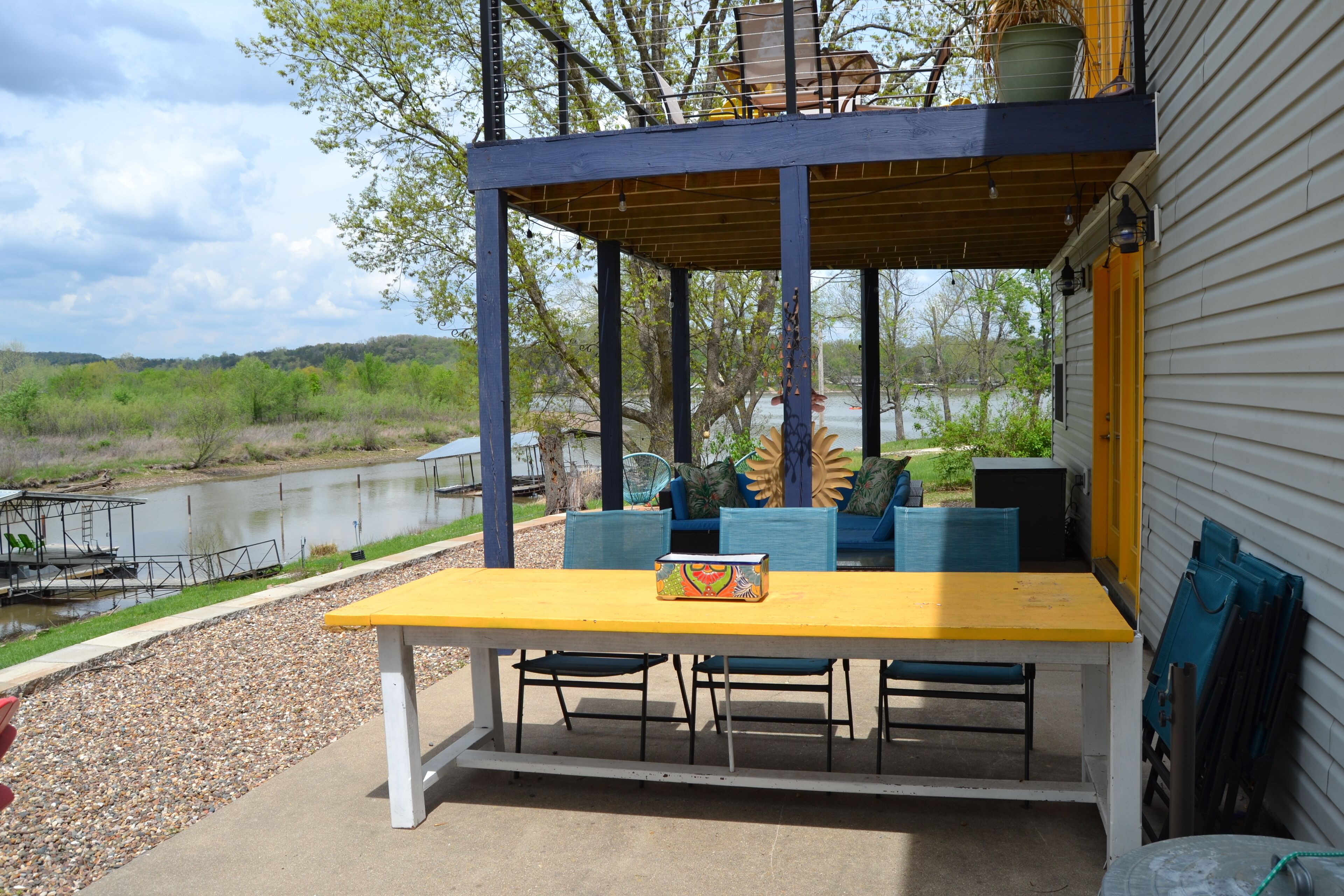 An outdoor patio with a yellow table and blue chairs, overlooking a body of water and surrounded by greenery.