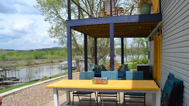 An outdoor patio with a yellow table and blue chairs, overlooking a body of water and surrounded by greenery.