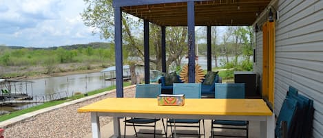 An outdoor patio with a yellow table and blue chairs, overlooking a body of water and surrounded by greenery.