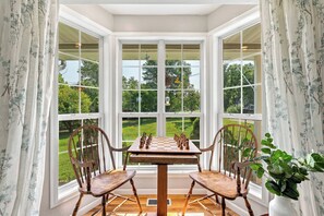 A bay window with a chessboard set up on a wooden table, flanked by two wooden chairs, overlooking a lush garden.