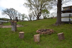 Un espacio al aire libre con césped, una fogata, troncos de madera y un árbol grande.