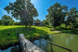 A house with a deck overlooking a pond, surrounded by lush greenery and trees.