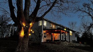 A house with a large tree in front, a covered porch with lights, and a clear sky.