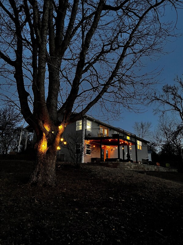 Una casa con un gran árbol en frente, un porche cubierto con luces y un cielo despejado.