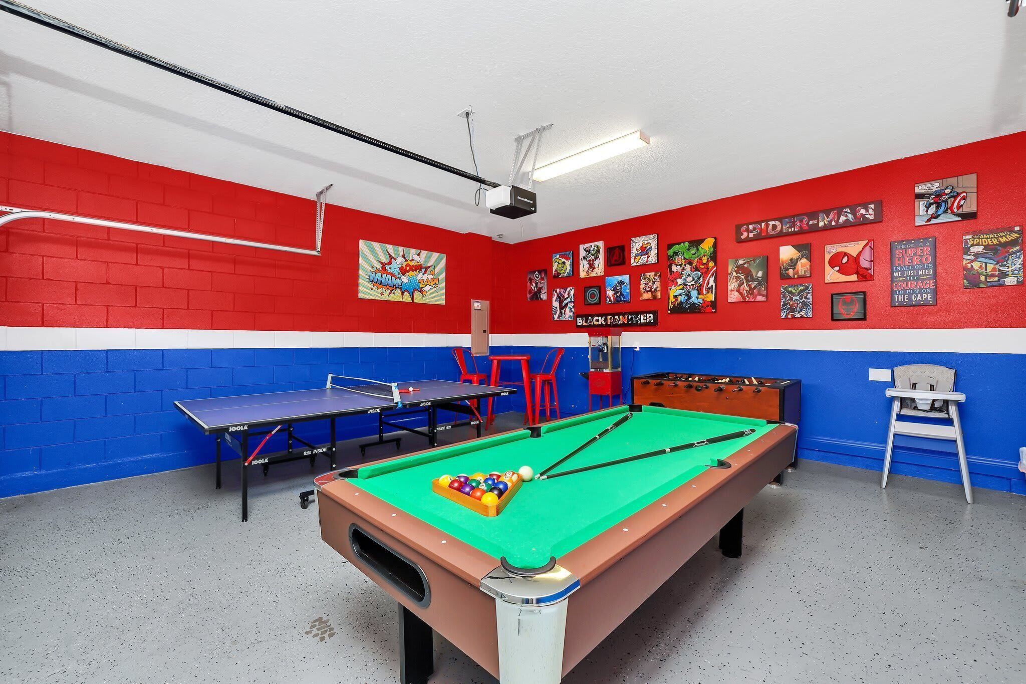 Une table de billard recouverte d’un drap de feutre vert, un mur rouge et bleu orné de posters de bandes dessinées, et une table de ping-pong à l’arrière-plan.